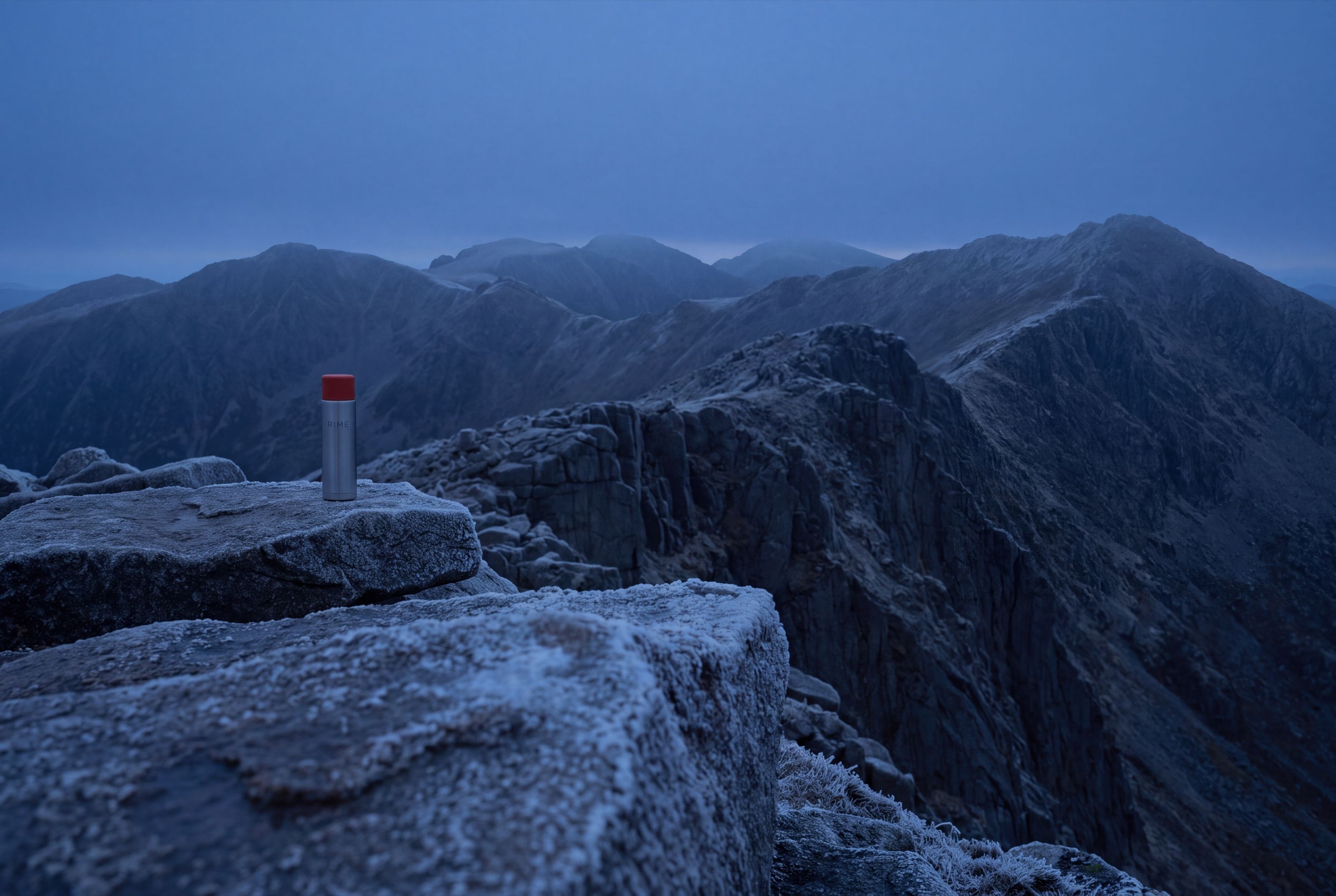 A steel RIME flask with an orange lid, small on a pre-dawn alpine ridge under deep blue light